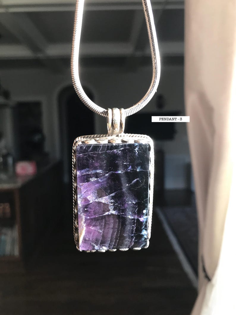 May include: A silver chain necklace with a rectangular pendant featuring a purple and blue fluorite stone. The pendant has a silver border with a scalloped edge. The text "PENDANT - 3" is visible in the image.