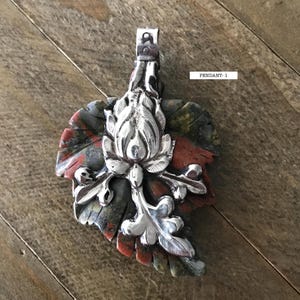 May include: A silver pendant with a leaf-shaped stone base, featuring red, green, and gray hues. The pendant has a detailed floral design in the center and the text "PENDANT-1".