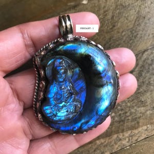 May include: A handcrafted pendant featuring a carved Buddha figure within a shimmering, iridescent blue and green stone. The pendant has a silver-colored metal bail and a decorative border. The text "PENDANT - 1" is visible.