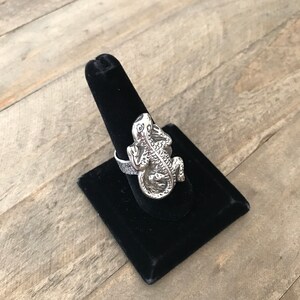 May include: A silver ring featuring a detailed lizard design. The ring is displayed on a black velvet ring holder.