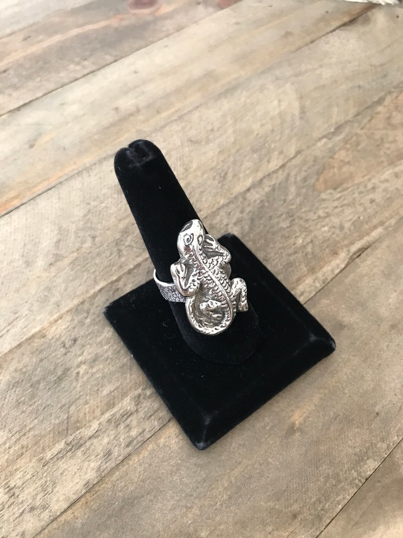 May include: A silver ring featuring a detailed lizard design. The ring is displayed on a black velvet ring holder.
