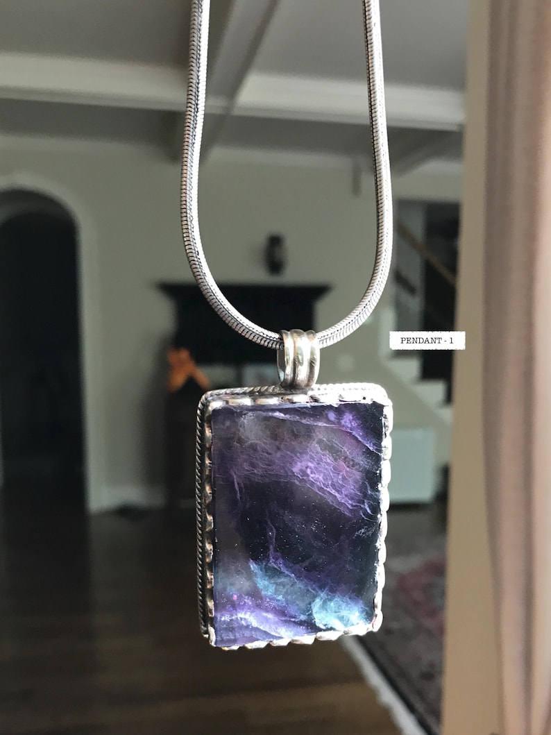 May include: A silver chain necklace with a rectangular pendant featuring a purple, blue, and green gemstone set in a silver frame. The pendant has a scalloped edge.