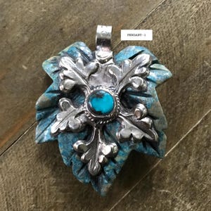 May include: A turquoise and silver pendant in the shape of a leaf. The pendant has a central turquoise stone surrounded by a silver floral design. The leaf is carved with intricate details, and the pendant is labeled "PENDANT - 1".