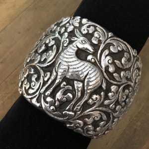 May include: A wide silver cuff bracelet featuring a detailed embossed relief of a standing deer surrounded by intricate swirling floral and vine patterns. The metal has a polished finish on the raised surfaces and a darkened, oxidized background.