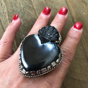 May include: A large, heart-shaped ring with a black, polished stone. The ring has a silver-colored, rope-like border and decorative accents. The ring is on a hand with red nail polish.