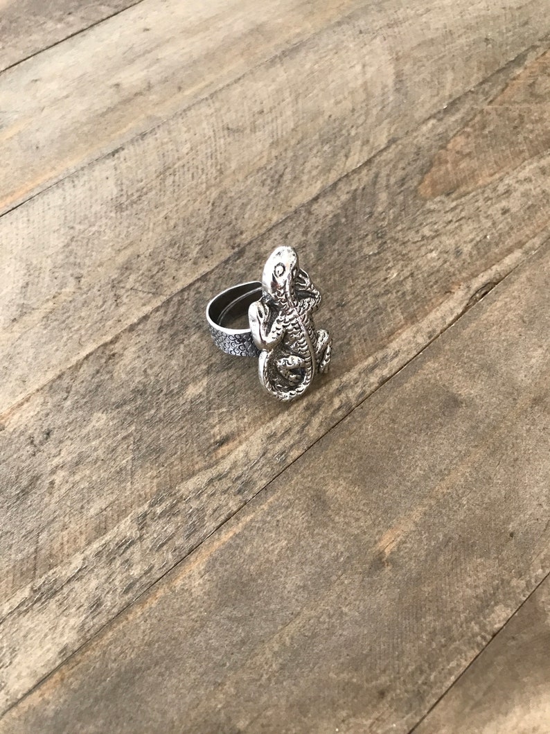 May include: A silver ring with a lizard design. The lizard is facing forward with its mouth open and its eyes closed. The ring is made of metal and has a smooth finish.