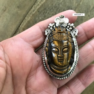 May include: Silver pendant featuring a carved tiger's eye stone with a Buddha face. The pendant has a floral design at the top and a beaded border.
