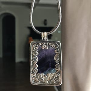 May include: A silver chain necklace with a rectangular pendant featuring a purple and green stone set in a silver frame with intricate floral designs.