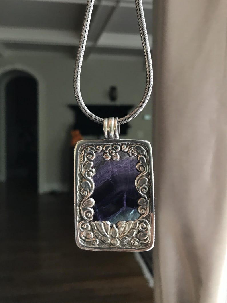 May include: A silver chain necklace with a rectangular pendant featuring a purple and green stone set in a silver frame with intricate floral designs.