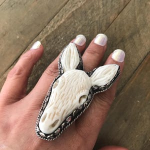 May include: A large, ivory-colored ring featuring a carved animal head design. The ring has a detailed, textured surface and is set in a silver-toned metal frame. The animal head has prominent ears and facial features.