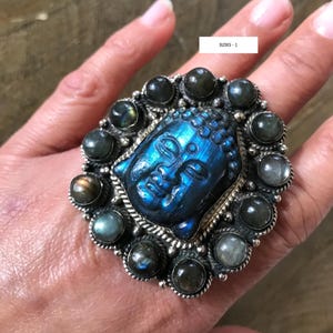 May include: A large silver ring featuring a carved blue Buddha face at the center, surrounded by a halo of dark, round gemstones. The ring is labeled "RING - 1".
