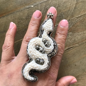May include: A large, ornate ring featuring a white snake design. The snake is intricately carved with a textured pattern and accented with silver-toned metal. The ring has a detailed, decorative border and a small floral element at the top.