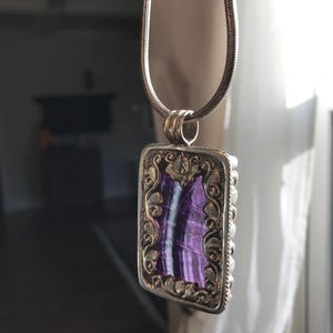 May include: A silver chain necklace with a rectangular pendant featuring a purple and white stone set in a gold-toned frame with intricate floral designs.