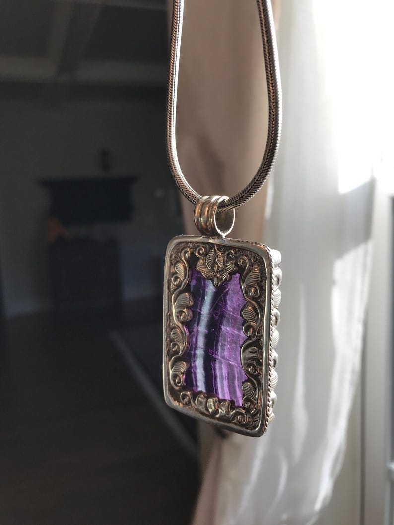 May include: A silver chain necklace with a rectangular pendant featuring a purple and white stone set in a gold-toned frame with intricate floral designs.