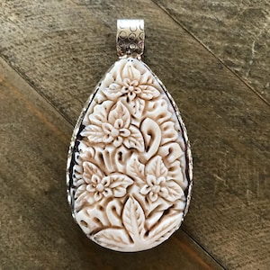 May include: Cream-colored pendant with a floral design carved into the surface. The pendant has a teardrop shape and is framed with a silver-colored metal. The bail is also silver-colored and has a decorative pattern.