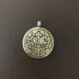 May include: A silver-toned pendant with an intricate design featuring a stylized flower and swirling patterns. The pendant is round and has a loop at the top for attaching a chain.