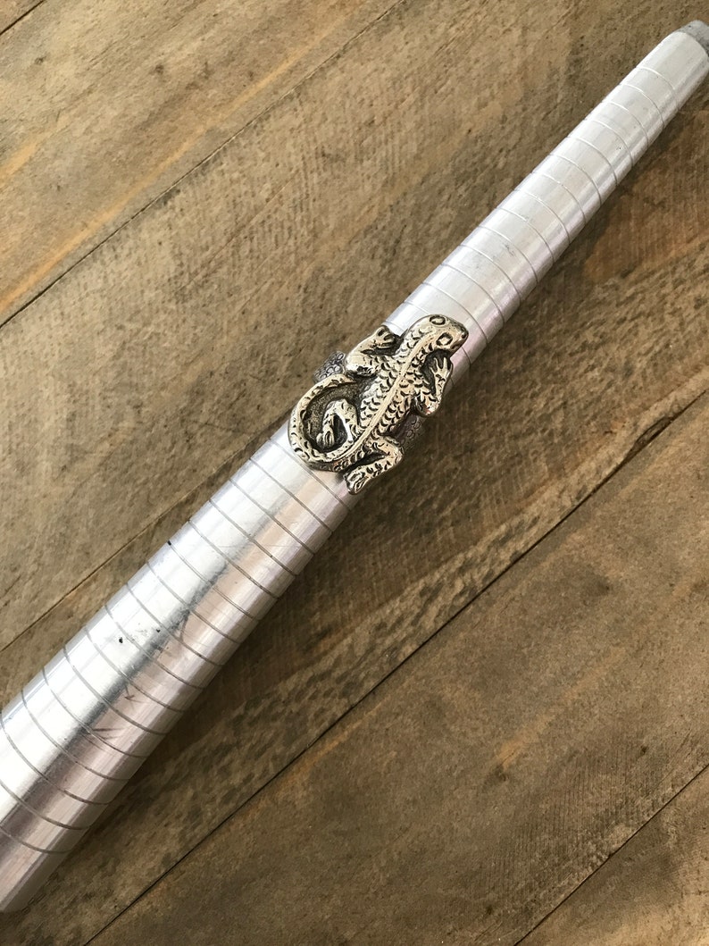May include: A silver lizard ring on a metal ring sizer. The ring is detailed with scales and has a textured finish.