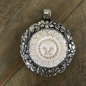May include: A silver pendant with a carved ivory-colored sun design. The sun has a serene face and is surrounded by a detailed floral and scrollwork border. The pendant has a bail for a chain.