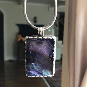 May include: A silver chain necklace with a rectangular pendant featuring a dark purple and blue iridescent glass stone.