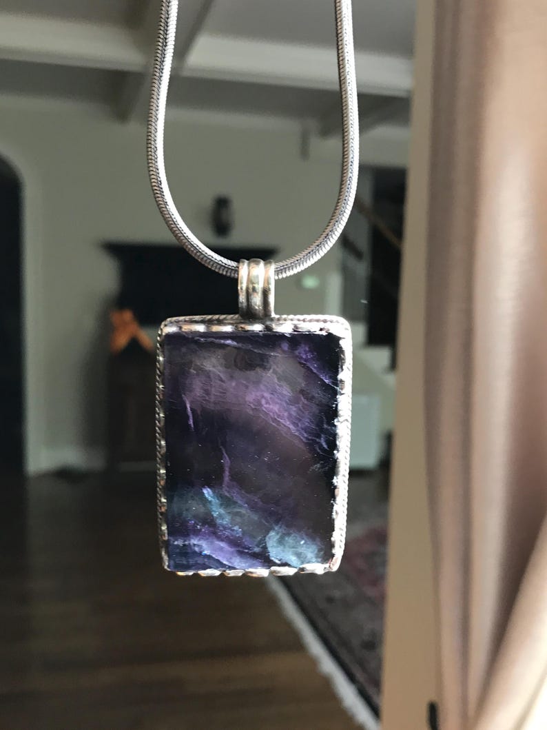May include: A silver chain necklace with a rectangular pendant featuring a dark purple and blue iridescent glass stone.
