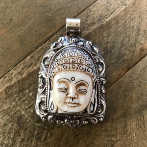 May include: A silver pendant featuring a carved Buddha head. The Buddha's face is cream-colored with detailed facial features and a serene expression. The pendant is framed by an ornate silver border, showcasing an antique aesthetic.