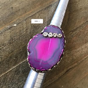 May include: A large, statement ring featuring a vibrant, heart-shaped, fuchsia agate stone. The stone is set in a silver-toned metal with a decorative border. The text "RING - 1" is visible.