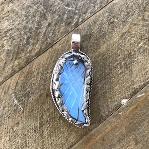 May include: A silver pendant with a light blue, leaf-shaped stone. The stone has a cross-hatched pattern and is set in a decorative silver frame. The pendant has a silver bail for a chain.