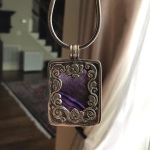 May include: A silver necklace with a rectangular pendant featuring a purple stone surrounded by an intricate floral design.