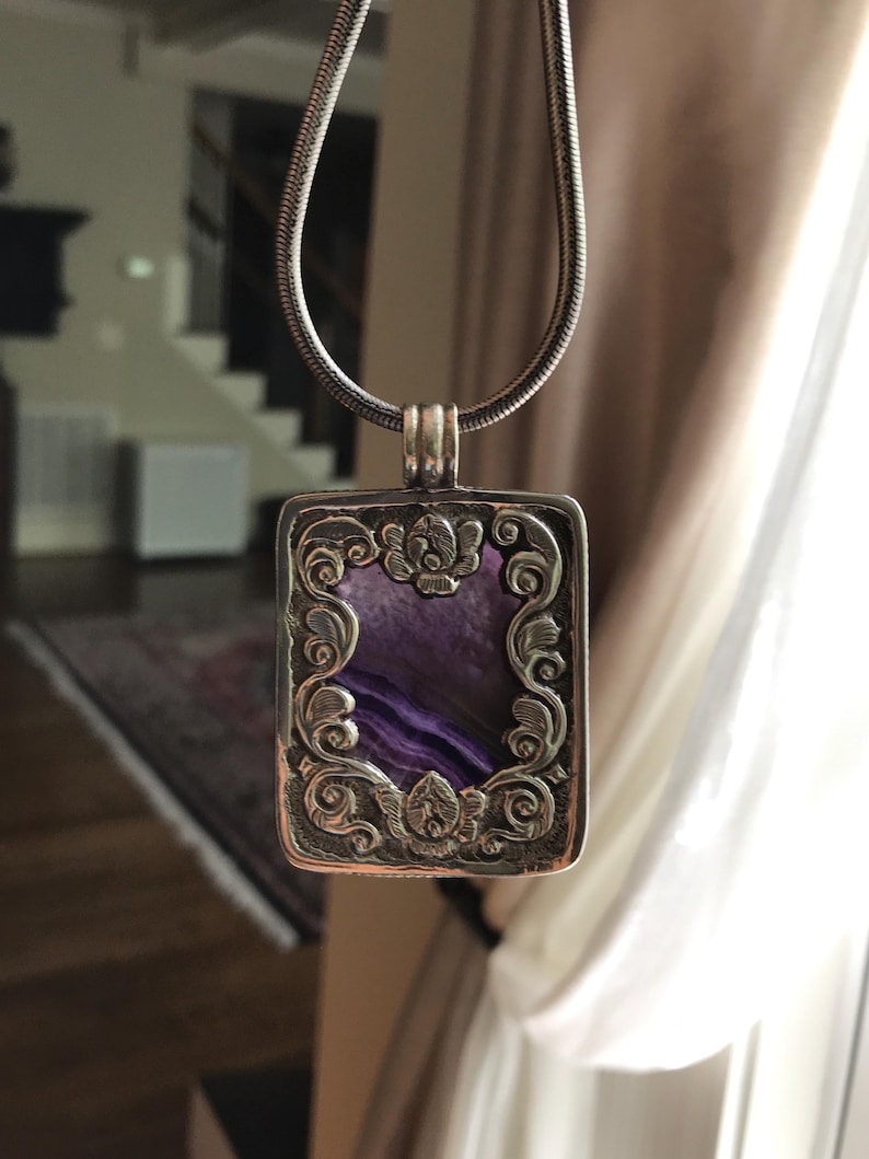 May include: A silver necklace with a rectangular pendant featuring a purple stone surrounded by an intricate floral design.