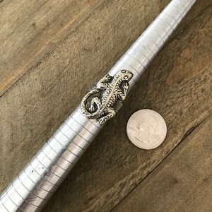 May include: A silver ring with a detailed lizard design. The ring is shown on a ring sizer with a US quarter for scale.