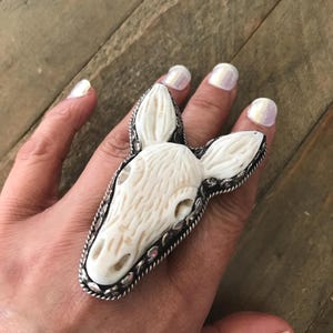 May include: A statement ring featuring a carved off-white animal head with pointed ears and textured details. The central carving is set within a dark, oxidized silver-toned metal frame that features a twisted rope border and scalloped edges.
