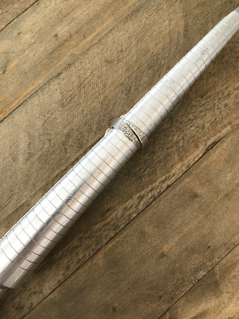 May include: A silver ring sizing tool with a silver ring on it. The tool is made of metal and has a series of grooves that allow you to measure the size of a ring.