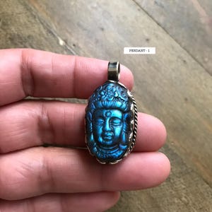 May include: A blue pendant featuring a carved Buddha head. The pendant has a silver-colored bail and a decorative silver-colored border. The text "PENDANT - 1" is visible in the image.