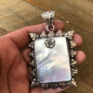 May include: A rectangular silver pendant with an iridescent mother-of-pearl center. The pendant has an ornate, filigree-style border and a small flower detail at the top. The bail is also intricately designed.