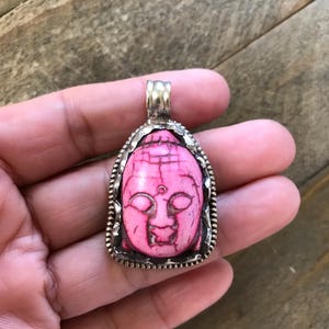 May include: A pink Buddha pendant with a silver-colored frame. The pendant features a detailed carving of a Buddha's face. The frame has a textured, beaded design. The pendant is a unique piece of jewelry.