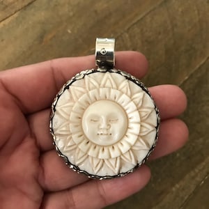 May include: A round, off-white pendant featuring a carved sun face with closed eyes, surrounded by a detailed sunburst design. The pendant is set in a silver-colored metal frame and has a small loop at the top for a chain.