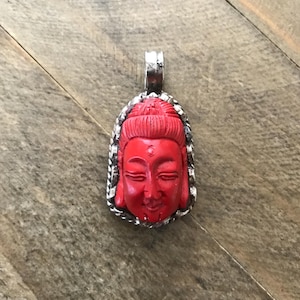 May include: A red carved pendant of a Buddha face with a silver frame.