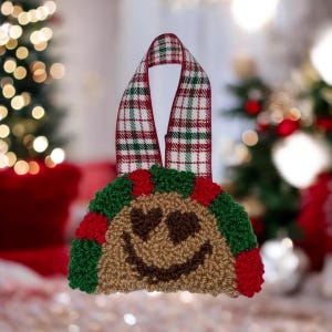 May include: A Christmas decoration shaped like a taco, with a smiling face and heart-shaped eyes. The ornament is made of textured brown, green, and red yarn, with a plaid ribbon for hanging. Festive holiday decor.
