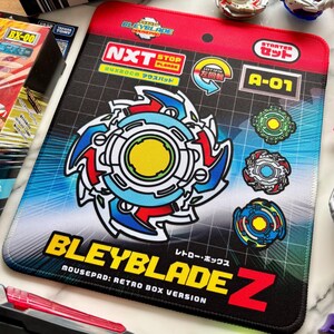 Op de afbeelding: Een rechthoekige Bleyblade Z-muismat met een groot, kleurrijk Beyblade-ontwerp in het midden. De muismat heeft de tekst "BLEYBLADE Z" en "MOUSEPAD: RETRO BOX VERSION". Er zijn extra afbeeldingen en tekst aanwezig, waaronder "NXT" en "A-01".