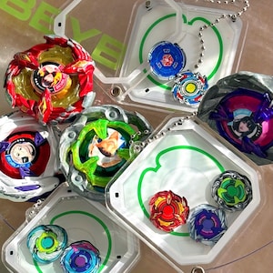 May include: A collection of Beyblade spinning tops in various colors including red, green, blue, and purple, featuring character portraits in the center. Several tops are stored inside clear plastic cases with green circular designs and metal chains.