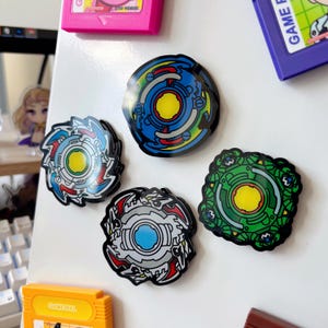 May include: Four colorful, circular magnets with intricate designs, resembling spinning tops. The magnets feature shades of blue, green, red, and yellow, with black outlines. Additional game cartridges are visible in the background.