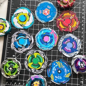 May include: A collection of circular Beyblade stickers featuring various colorful designs, including blue, red, green, and purple patterns. The stickers are arranged on a black cutting mat with white grid lines and measurement markings.