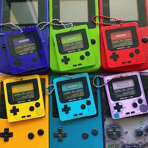 Può includere: Una collezione di portachiavi Game Boy Color colorati, tra cui blu, verde, rosso, giallo, ciano e viola. Ogni portachiavi presenta un design Game Boy in miniatura con schermo e pulsanti, con il testo "GAME PAL COLOR".