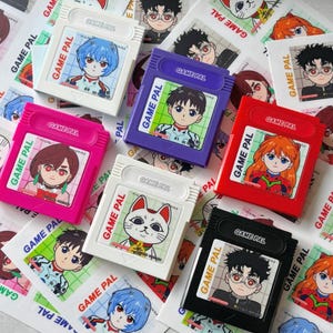 Könnte beinhalten: Eine Sammlung farbenfroher Game Pal-Cartridges mit Illustrationen von Anime-Charakteren. Die Cartridges sind in verschiedenen Farben erhältlich, darunter Weiß, Pink, Lila, Rot und Schwarz. Jede Cartridge hat ein anderes Charakterbild und den Text "GAME PAL".