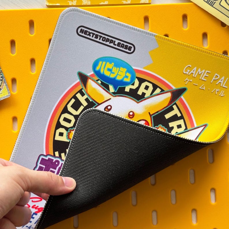 Op de afbeelding: Een grijs en gele muismat met een Pikachu-ontwerp, Japanse tekst en de woorden "GAME PAL" en "NEXTSTOPPLEASE". De muismat ligt op een gele pegboard-oppervlak. De zwarte, getextureerde onderkant is zichtbaar.