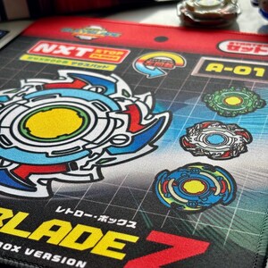 Op de afbeelding: Een kleurrijke speelmat met een gedetailleerde illustratie van een tol met blauwe, rode en gele accenten. De mat bevat Japanse tekst en afbeeldingen van andere toppen. Ontworpen voor een retro box versie. Afmetingen niet gespecificeerd.