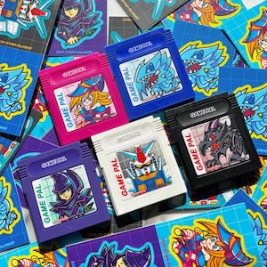 May include: A collection of colorful Game Pal video game cartridges. The cartridges are in various colors, including pink, blue, purple, white, and black. Each cartridge features a different anime-style character or robot illustration. The text "GAME PAL" is printed on each cartridge.