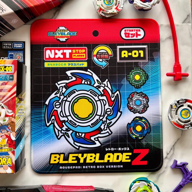 Op de afbeelding: Een Bleyblade-muismat met een blauw, rood en wit ontwerp, met de tekst "BLEYBLADE Z" en "MOUSEPAD: RETRO BOX VERSION." De muismat is 24 x 20 cm. Er zijn ook verschillende Bleyblade-tops en accessoires te zien.
