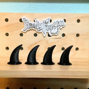 Op de afbeelding: Vier zwarte plastic voorwerpen in de vorm van haaienvinnen op een houten plank. De plank heeft een patroon van gelijkmatig verdeelde gaten. De tekst "Ready Fur You" is zichtbaar op een sticker achter de voorwerpen.