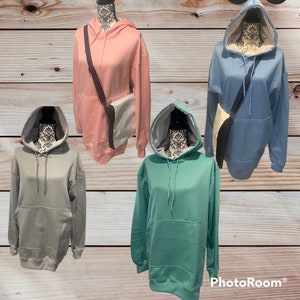 Op de afbeelding: Vier hoodies in verschillende kleuren: roze, lichtblauw, grijs en teal. Elke hoodie heeft een zak met een kangoeroe-stijl en een capuchon met een trekkoord. De hoodies worden getoond op mannequins met een bruine houten achtergrond.
